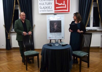 Janusz L. Wisniewski i prowadząca spotkanie Beata Przybylska siedzący przy małym okrągłym stoliku. Za stołem na sztalusze plakat promujący wydarzenie.